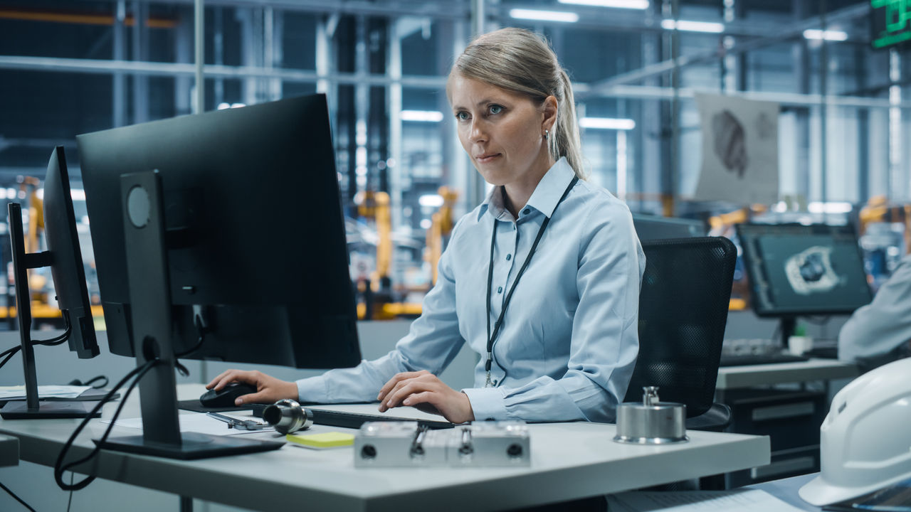 Car Factory Office: Portrait of Confident Female Chief Engineer Working on Desktop Computer. Professional Technician in Automated Robot Arm Assembly Line Manufacturing High-Tech Electric Vehicles
