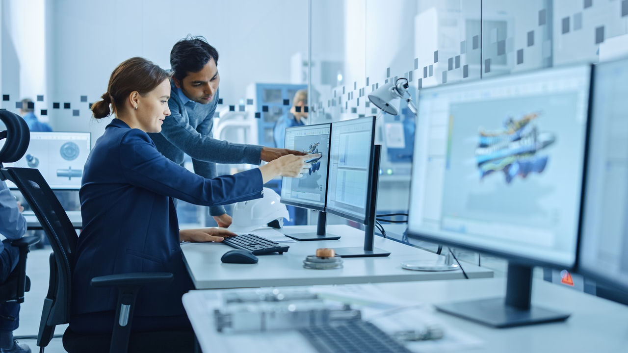 Inside Factory Office: Male Project Supervisor Talks to a Female Industrial Engineer who Works on Computer, Talk. In Workshop: Professional Workers Use High-Tech Industry 4 CNC Machinery, Robot Arm.